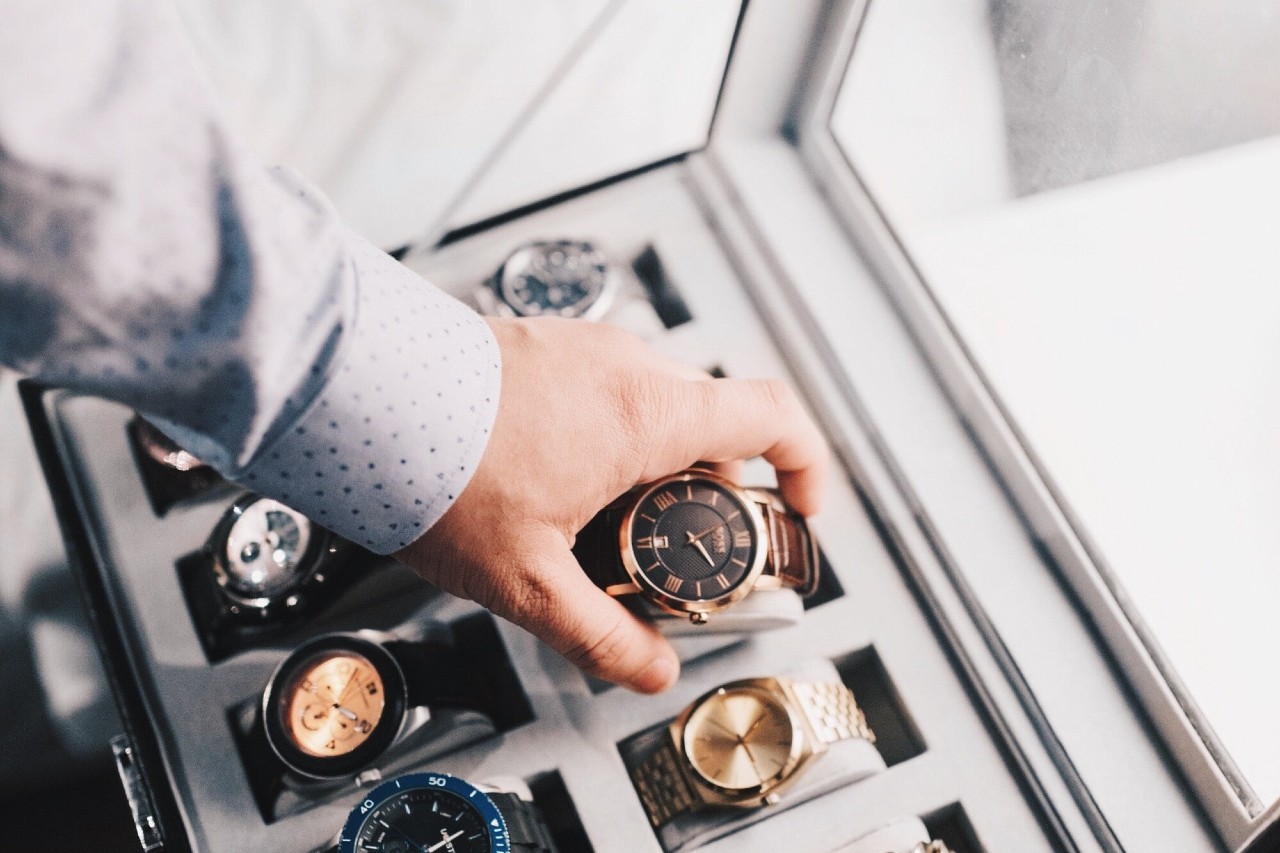 A hand in a polka dot shirt selects a brown watch from a display case filled with various elegant watches.