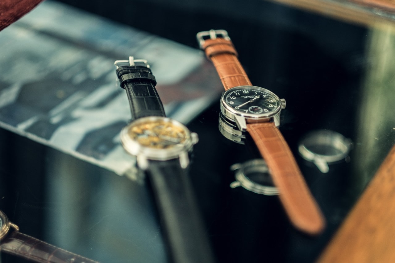 Two wristwatches with leather straps displayed on a glass surface.