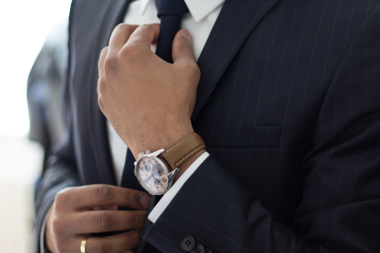 A man in a grey suit fixes his tie, showcasing a brown leather wristwatch and a yellow gold wedding band