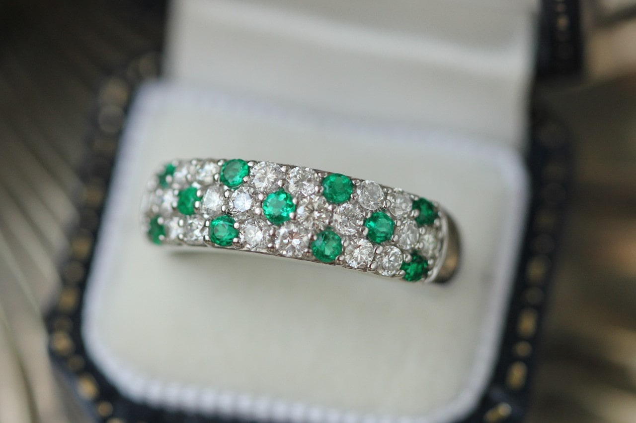 A close-up of a white gold ring with alternating rows of round brilliant cut diamonds and emeralds in an open ring box with a dark blue interior.