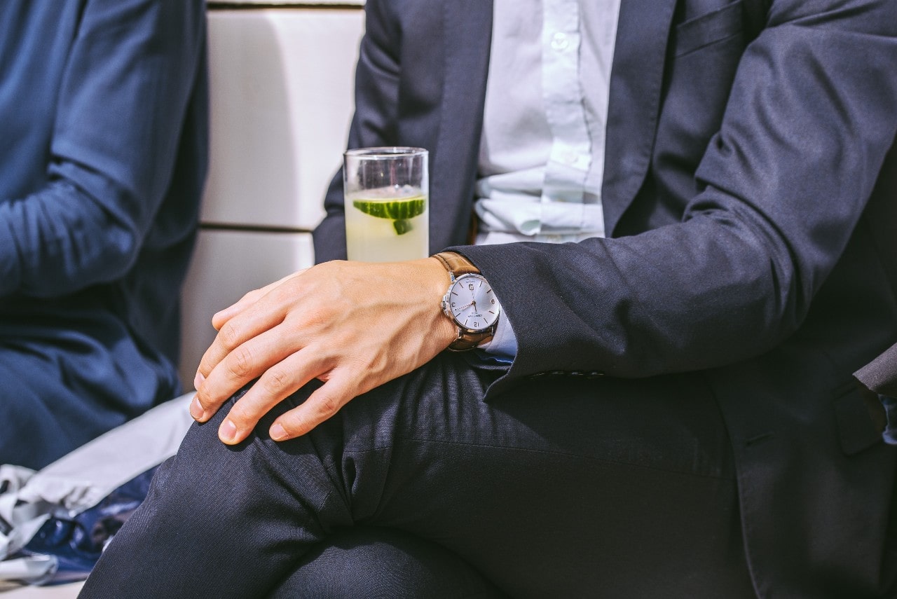 A man dressed in a navy suit and holding a drink wears a silver dress watch with a brown leather band.