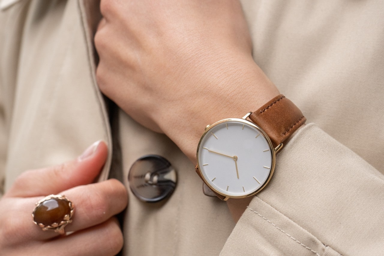 A close-up of a person wearing a minimalist gold watch with a tan leather strap, styled with a beige trench coat and a large oval ring.