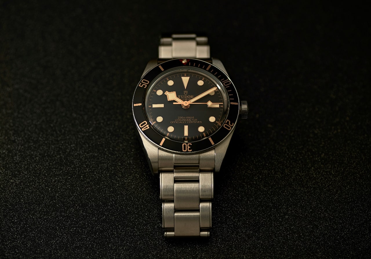 A TUDOR metallic watch with black dial on a black textured surface