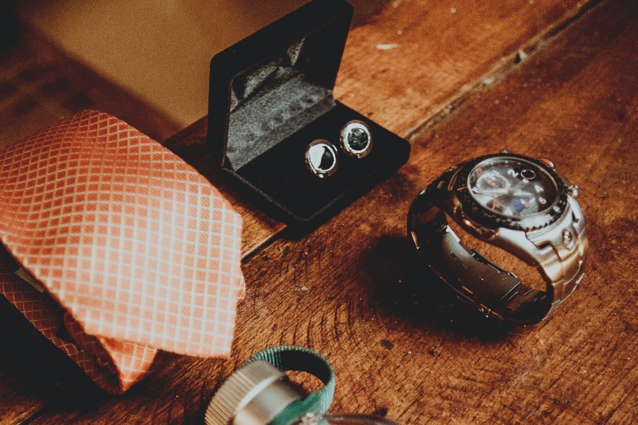Elegant men's accessories on a wooden surface: an orange patterned tie, cufflinks in a black case, and a silver watch.