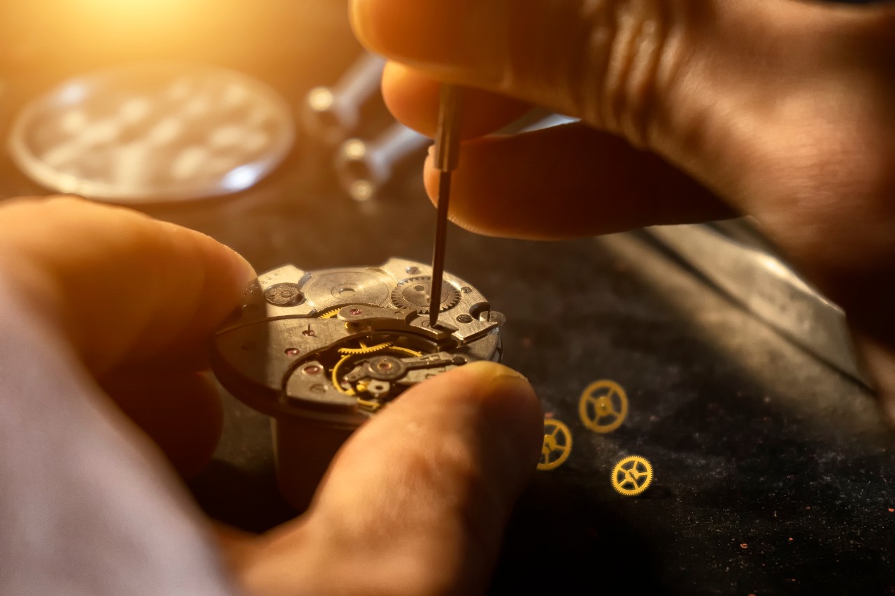 Close-up of a watchmaker&rsquo;s hands using a small screwdriver to repair a watch movement, with visible gears and cogs, warmly lit.