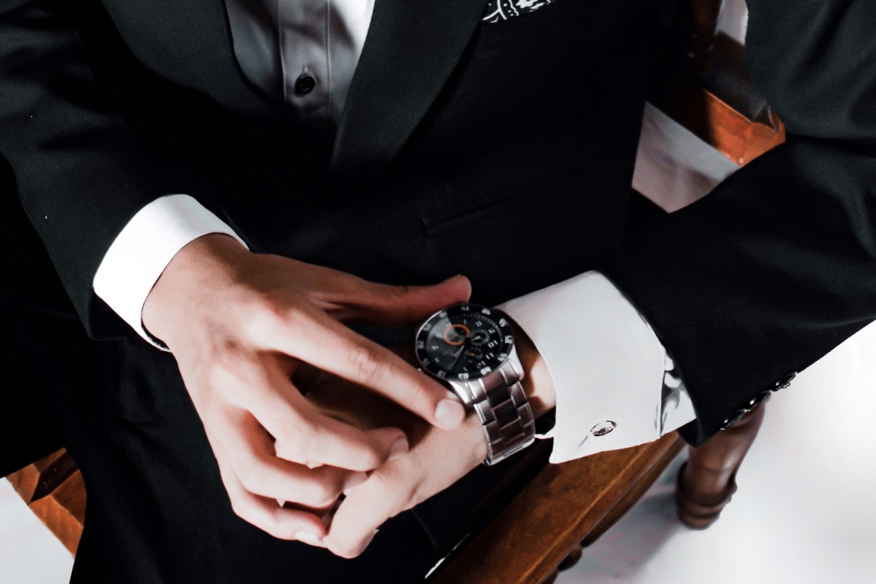 A man in a formal black suit adjusts a silver wristwatch, seated on a wooden chair.
