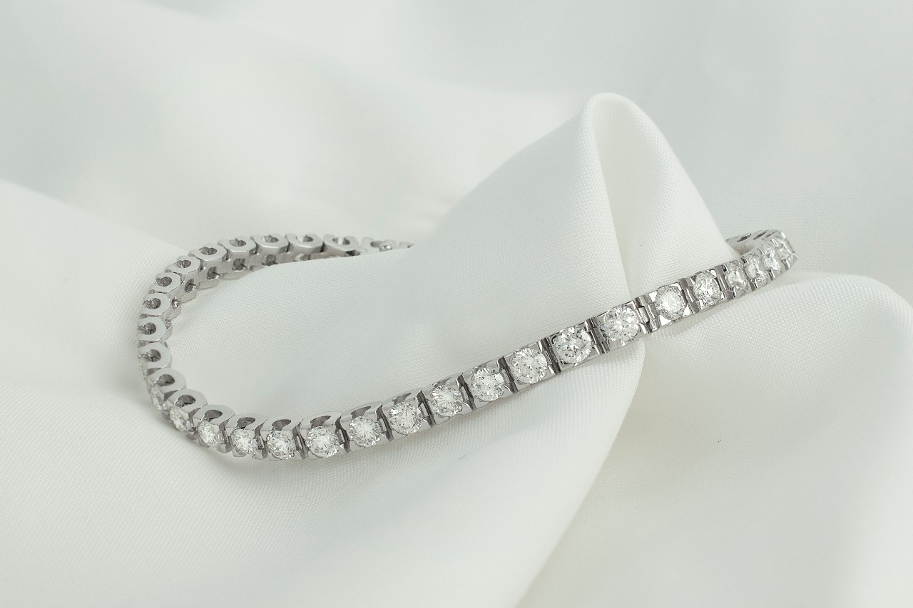 A diamond tennis bracelet rests on smooth white fabric, showcasing round diamonds set in a delicate continuous line.