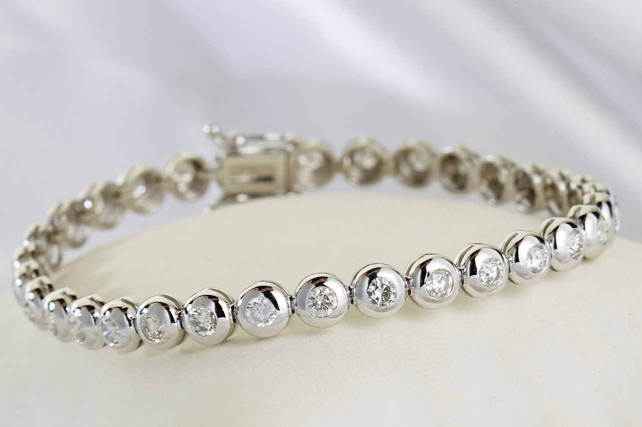 A silver tennis bracelet featuring round, sparkling diamonds set in bezel settings, resting on a smooth cream surface.