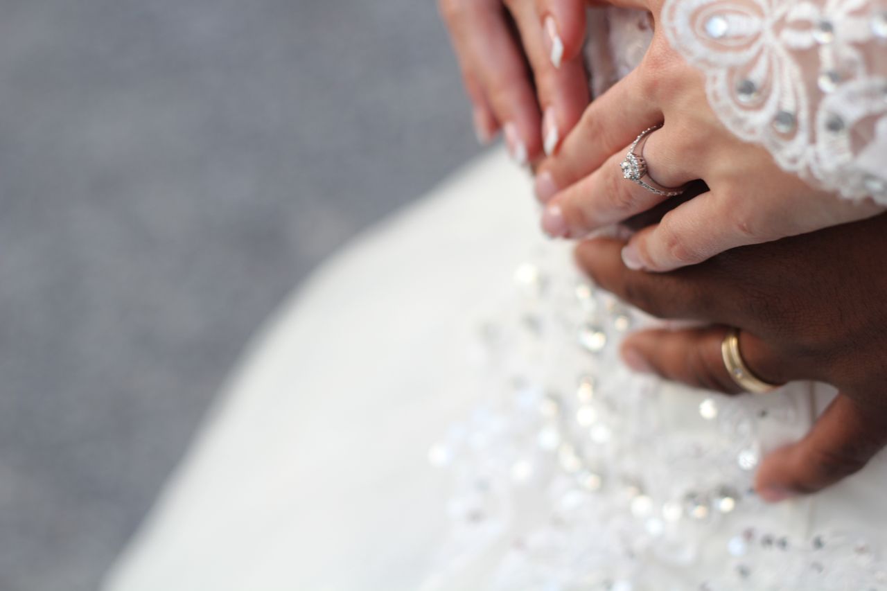 Close-up of intertwined hands, showcasing a bride's lace sleeve, engagement ring, and groom's wedding band.