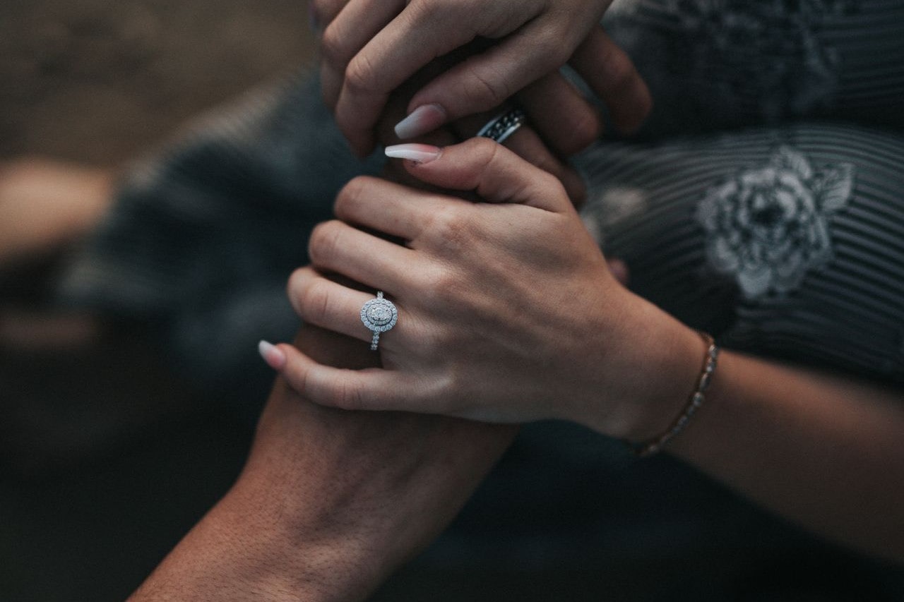 A woman holds a man’s hands wearing a halo engagement ring. A woman holds a man’s hands wearing a halo engagement ring.