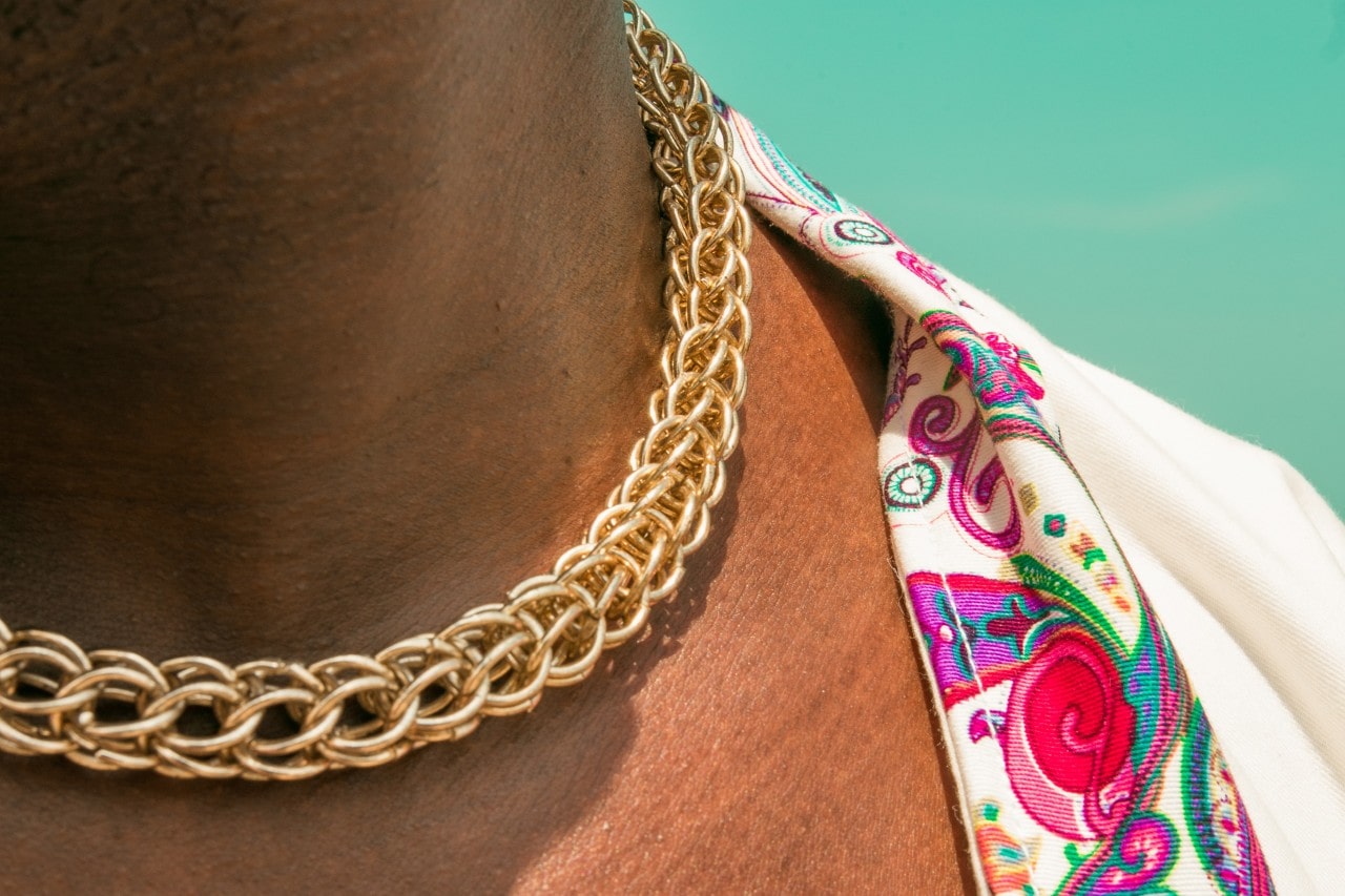 A close-up of a person wearing a thick gold chain necklace and a colorful paisley-patterned shirt against a turquoise background. A close-up of a person wearing a thick gold chain necklace and a colorful paisley-patterned shirt against a turquoise background.