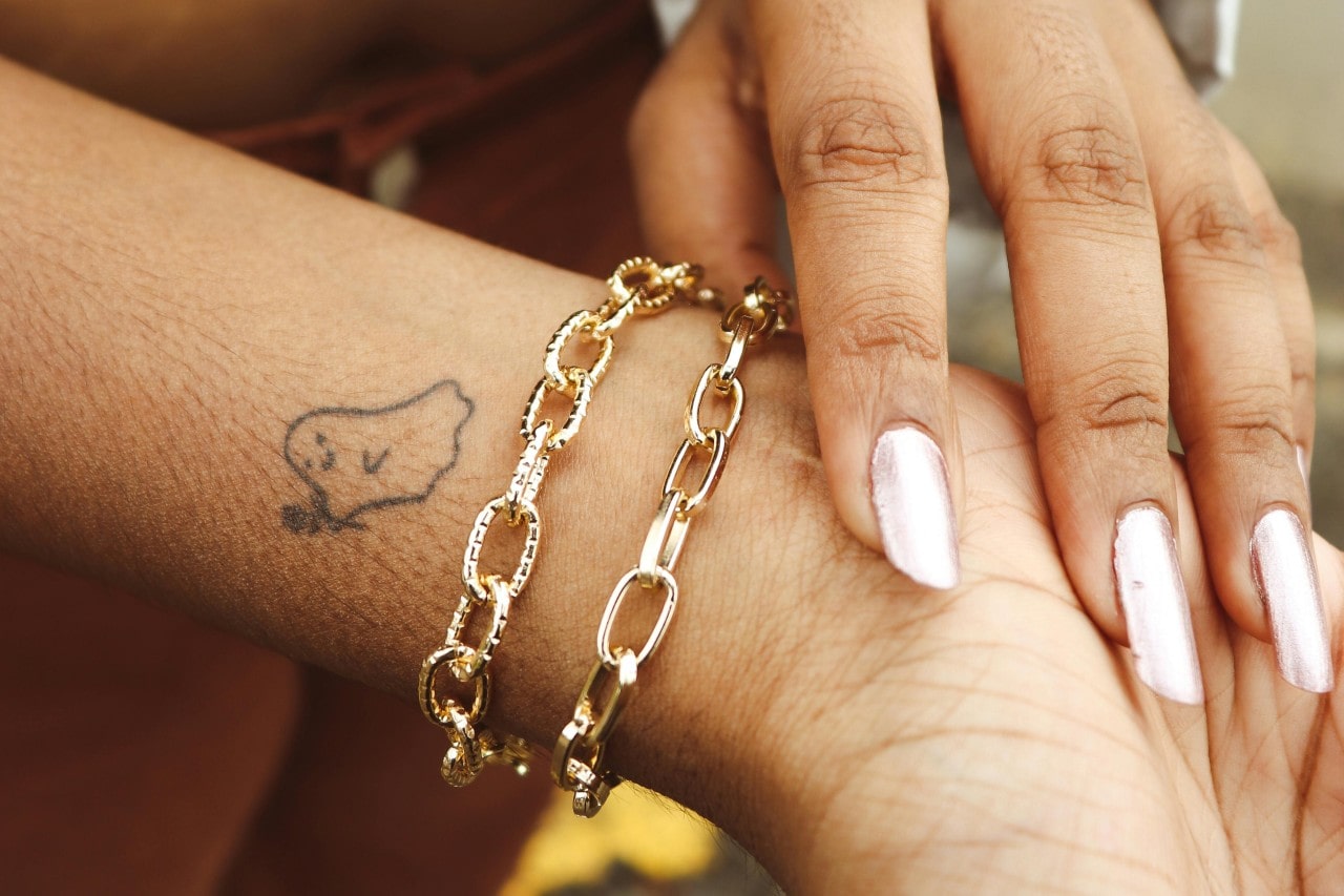 A close-up of two gold chain bracelets worn on a wrist with a small ghost tattoo. A close-up of two gold chain bracelets worn on a wrist with a small ghost tattoo.