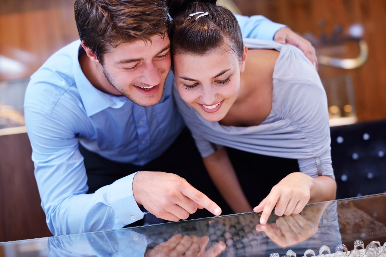 A smiling couple in a jewelry store leans over a glass display case, excitedly pointing at items.
