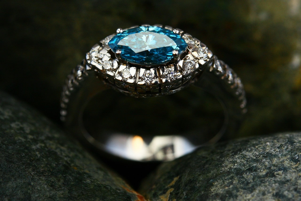 A silver halo ring with a blue gemstone at the center, surrounded by sparkling diamonds, resting on a dark, textured stone surface.