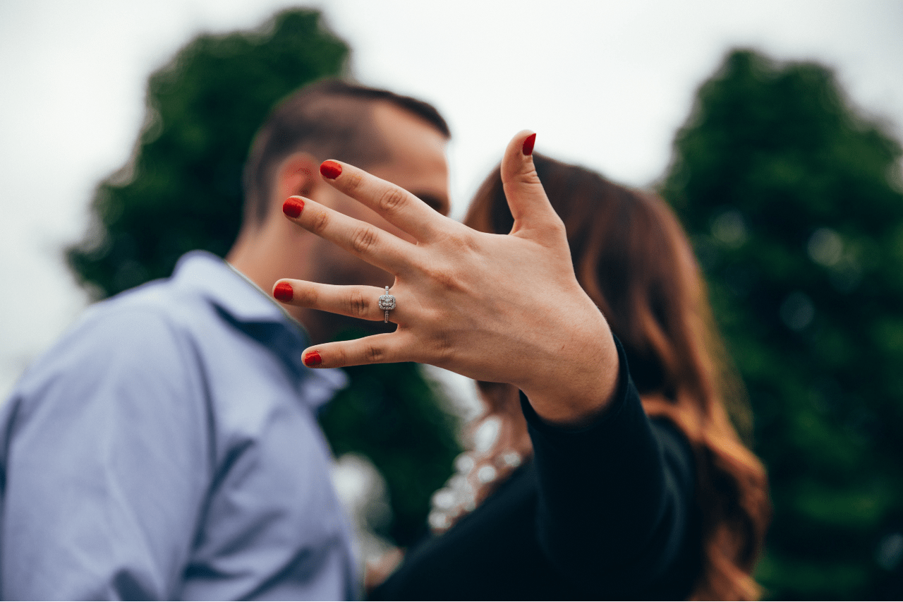 A woman's hand with red nails showcases a sparkling engagement ring, while she kisses a man in a blue shirt against a lush, blurred greenery.