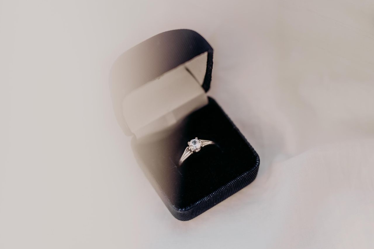 A classic solitaire engagement ring displayed in an open black velvet box, placed on a soft white surface. A classic solitaire engagement ring displayed in an open black velvet box, placed on a soft white surface.