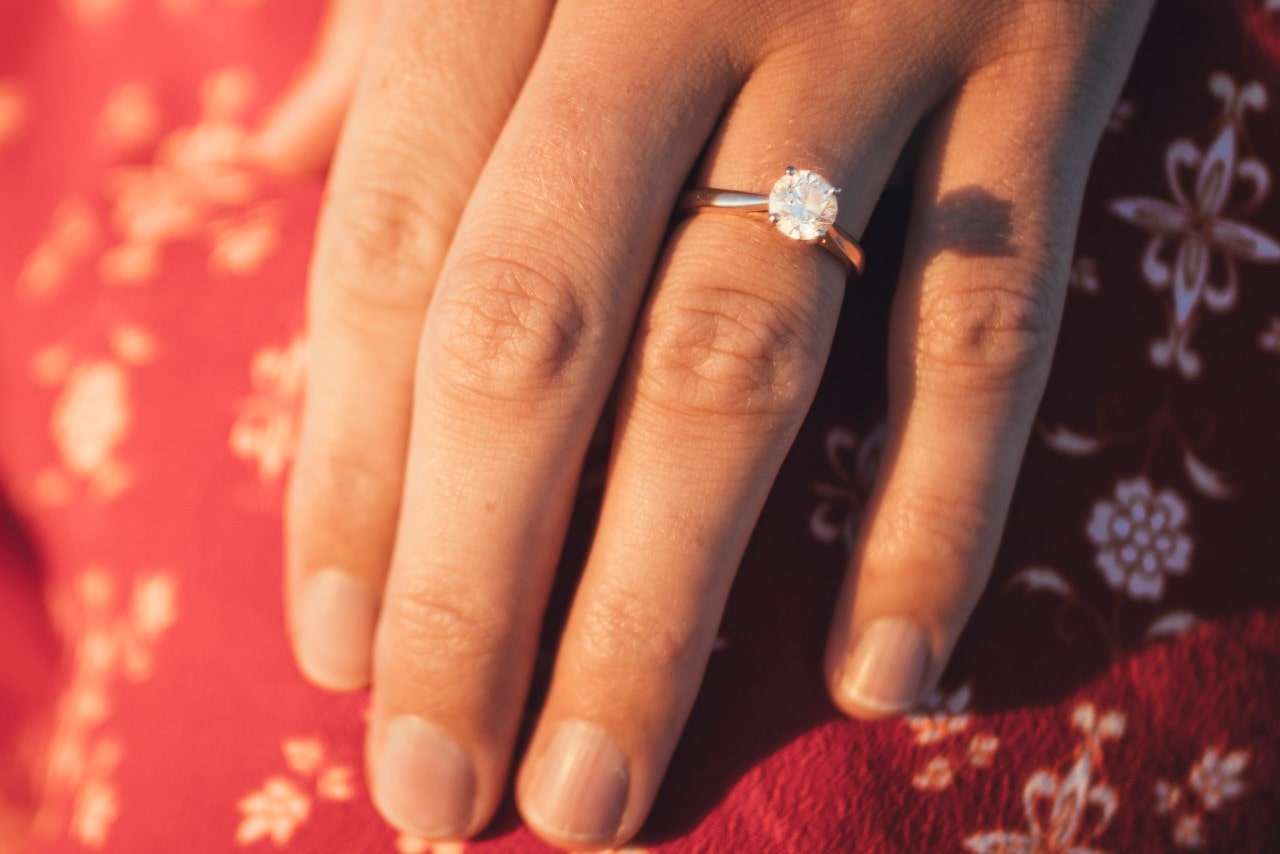 A close-up of a woman's hand wearing a solitaire engagement ring on her ring finger, with a red patterned fabric in the background. A close-up of a woman's hand wearing a solitaire engagement ring on her ring finger, with a red patterned fabric in the background.