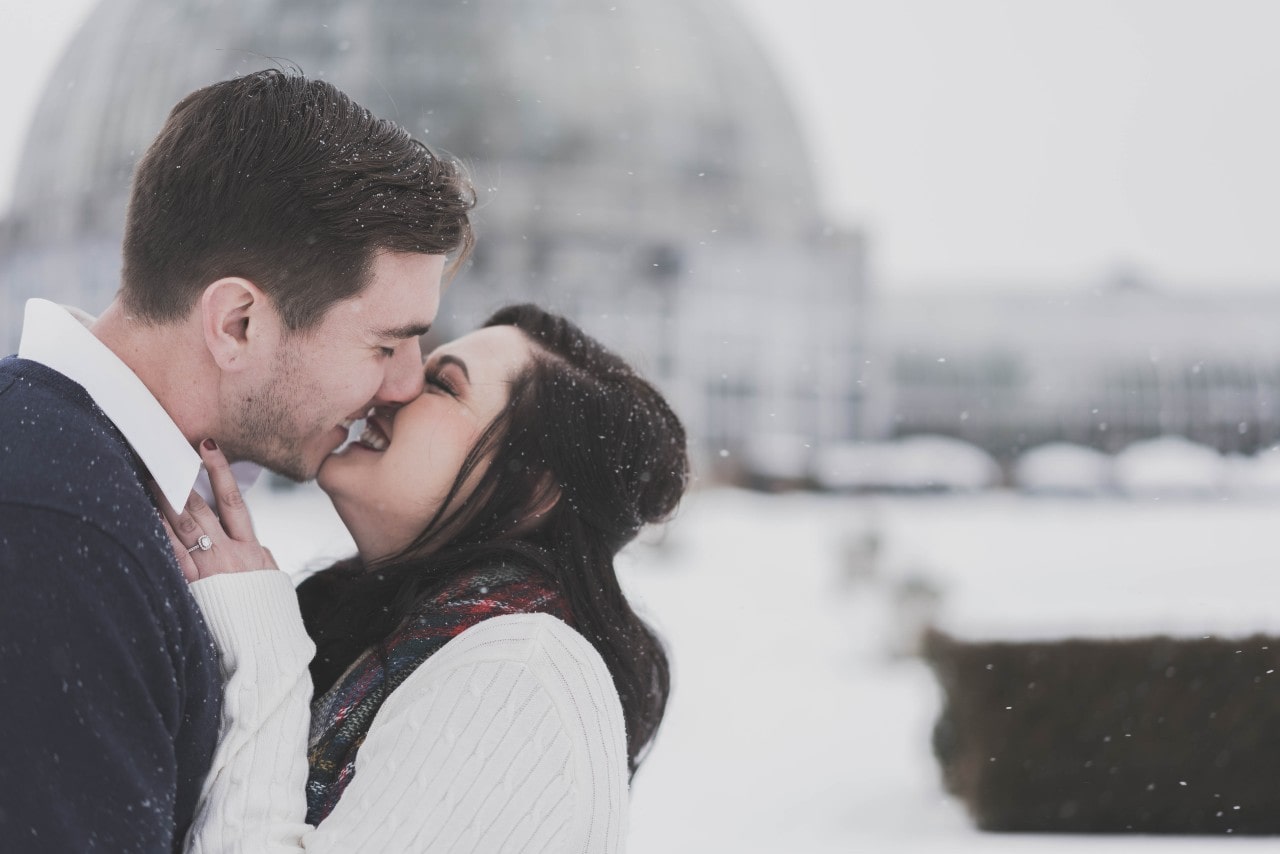 A happy couple embraces in the snow, smiling as they lean in for a kiss, with a domed building blurred in the background. A happy couple embraces in the snow, smiling as they lean in for a kiss, with a domed building blurred in the background.