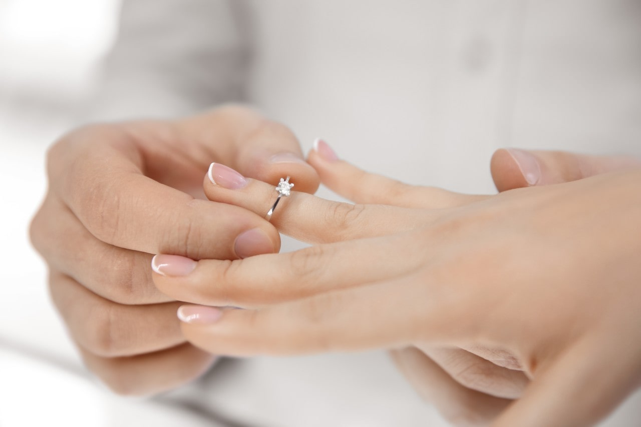 A person placing a white gold solitaire engagement ring on their partner's finger. A person placing a white gold solitaire engagement ring on their partner's finger.