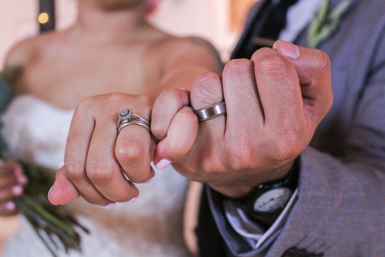 Close-up of a couple linking pinkies, both wearing wedding rings; the bride in a white dress holding a bouquet, the groom in a suit with a visible wristwatch. Close-up of a couple linking pinkies, both wearing wedding rings; the bride in a white dress holding a bouquet, the groom in a suit with a visible wristwatch.
