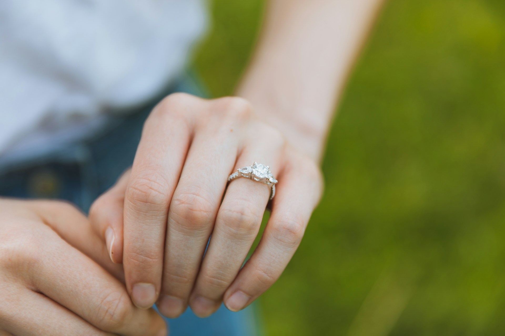A close up of a three stone diamond engagement ring on a woman’s hand against a blurry grass background A close up of a three stone diamond engagement ring on a woman’s hand against a blurry grass background
