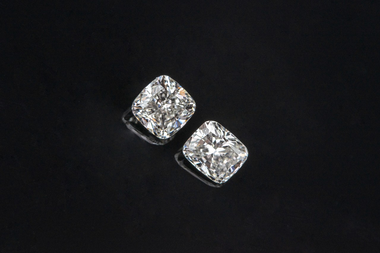 Two rounded square loose diamonds laying on a black surface, with slight reflect under each one.