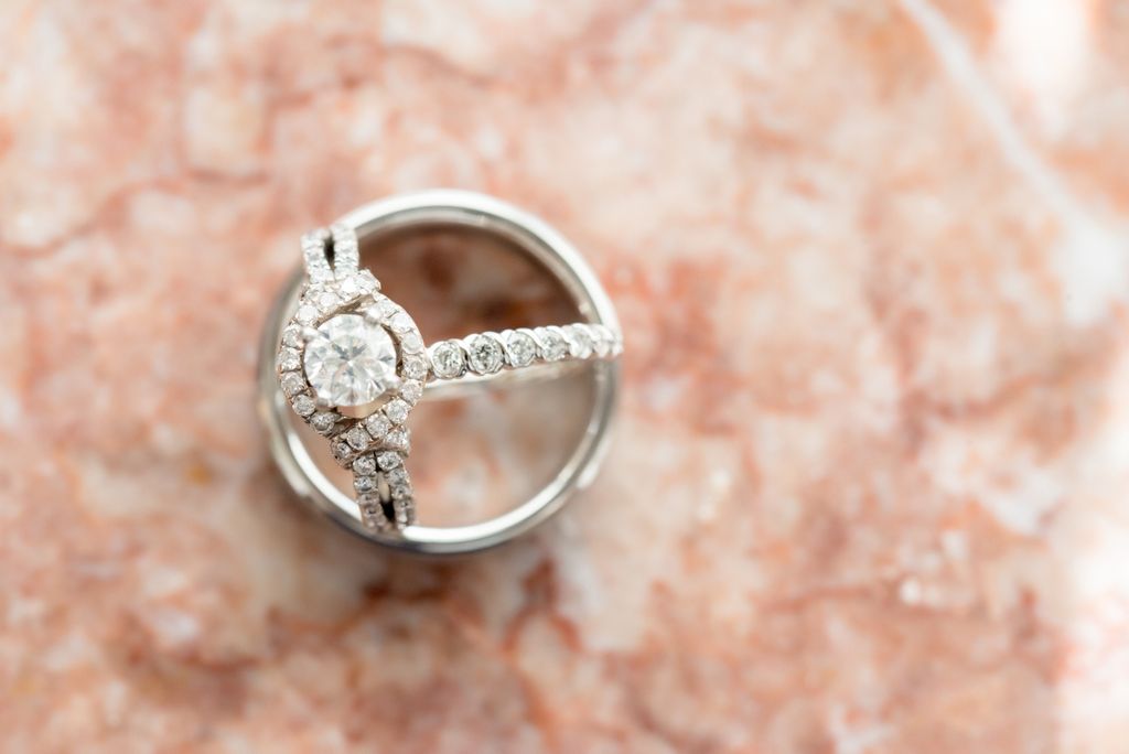 A diamond engagement ring with a circular diamond and a halo, showcased on a pink marble table.
