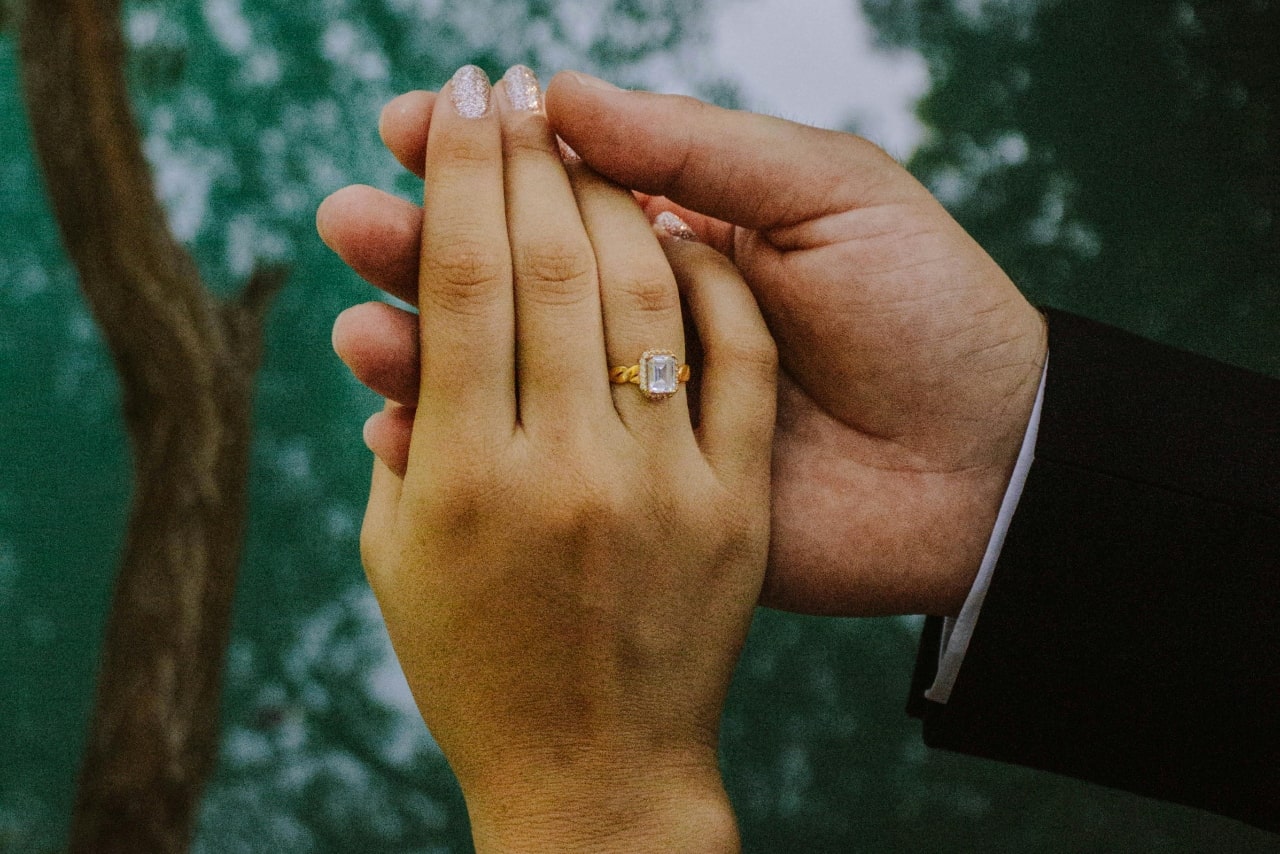 Two hands held together against a forestry landscape, showcasing a yellow gold emerald cut diamond engagement ring.