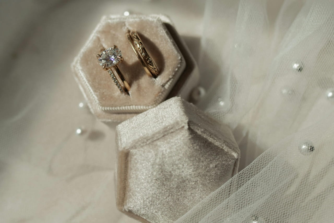 Two gold diamond wedding rings, one with a solitaire diamond, rest inside a soft velvet ring box on sheer fabric decorated with pearls.