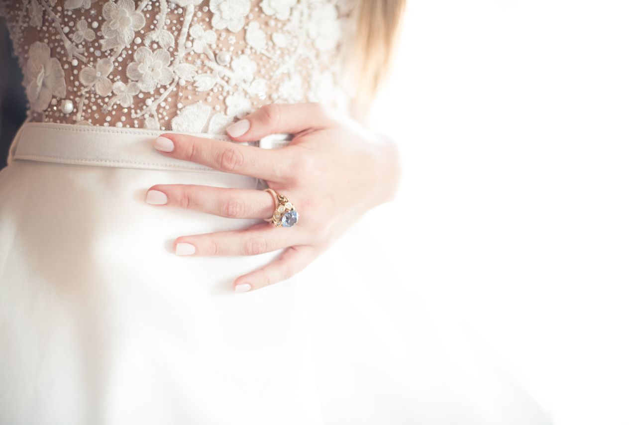 A bride’s hand with a gold ring featuring a vibrant gemstone, resting on her lace wedding gown A bride’s hand with a gold ring featuring a vibrant gemstone, resting on her lace wedding gown